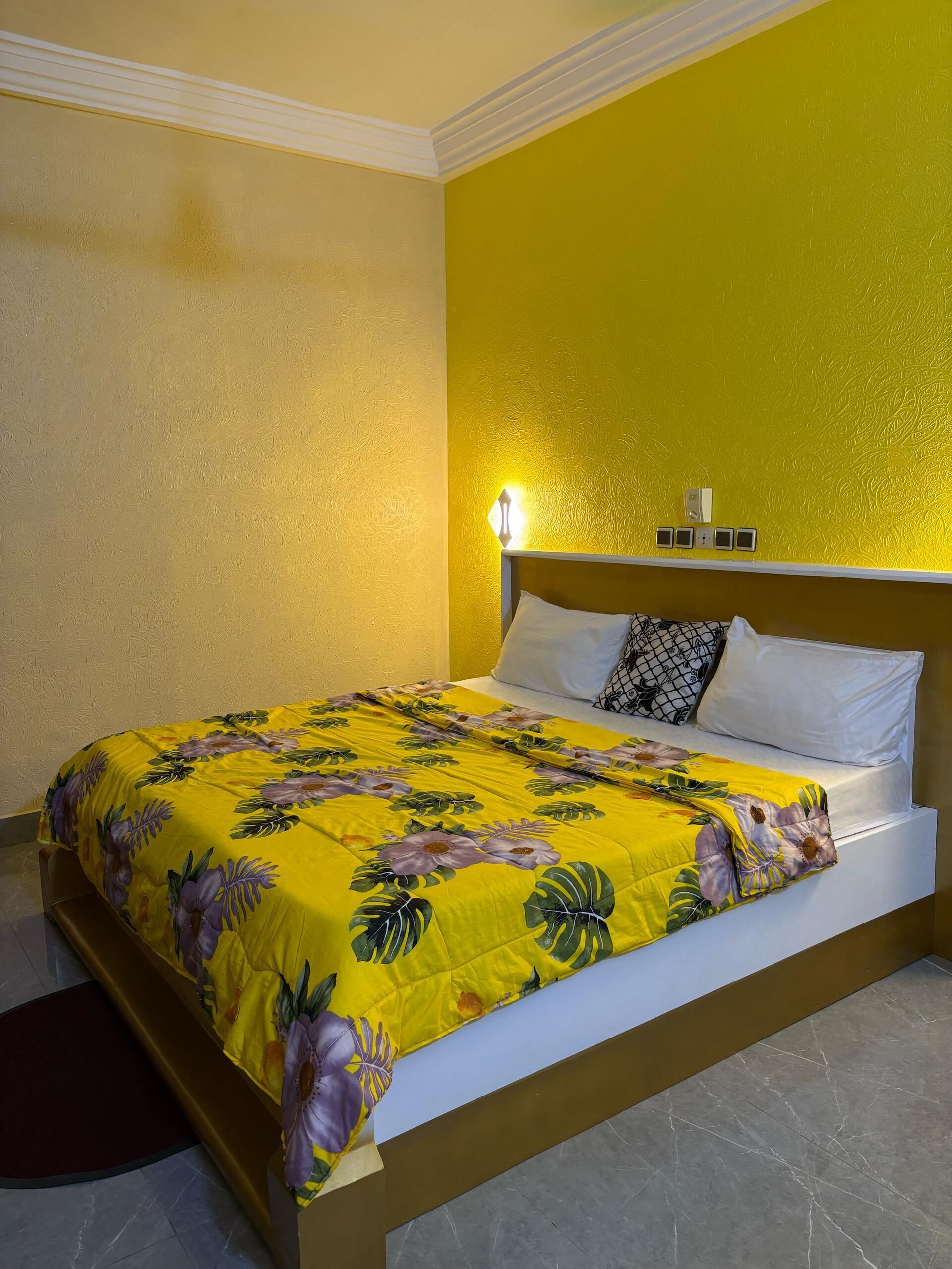 A hotel room with a bed, a nightstand, and a wall-mounted light.