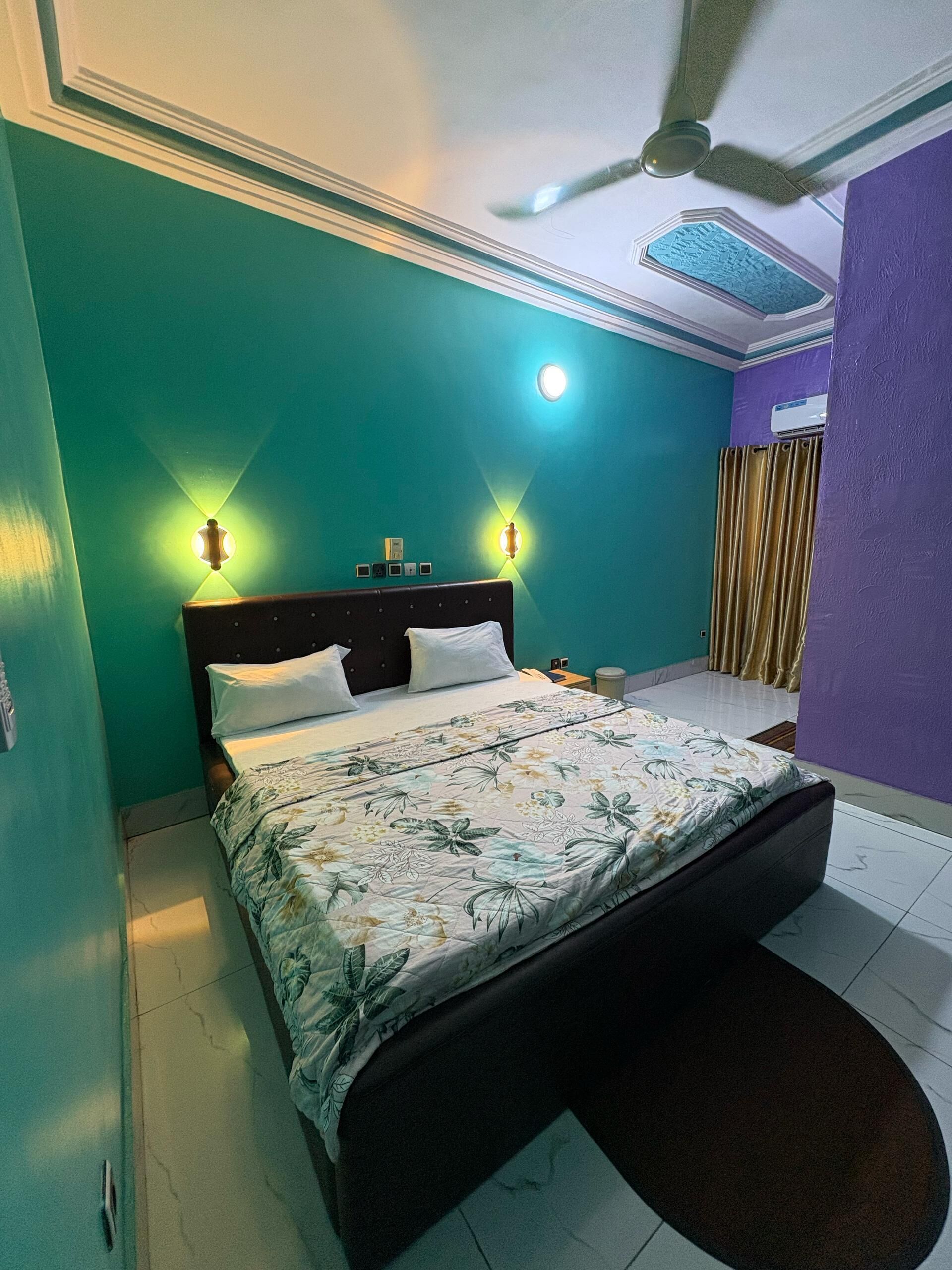 A bedroom with teal walls, a floral bedspread, a dark headboard, two wall-mounted lights, a ceiling fan, and an air conditioning unit.