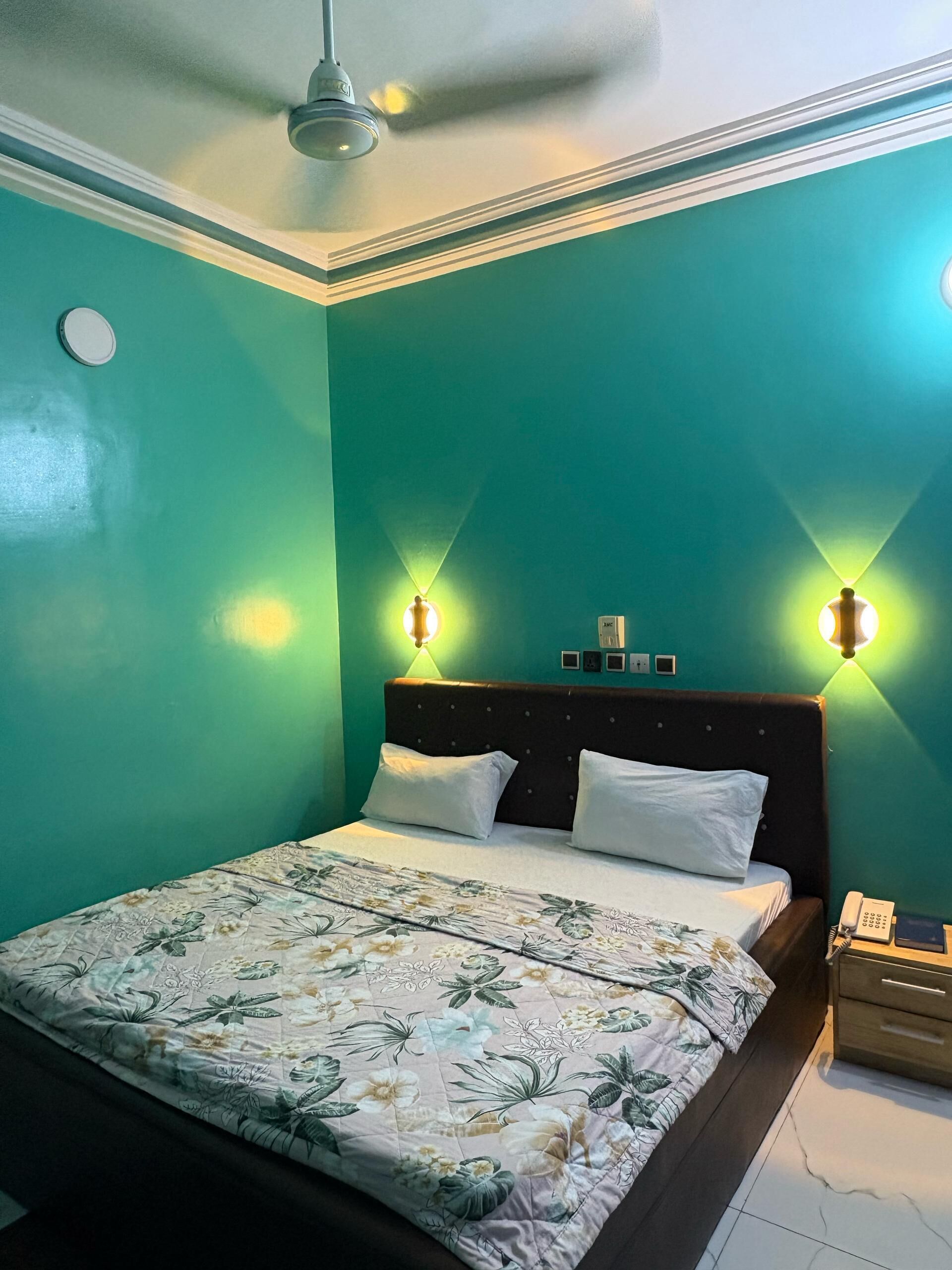 A bedroom with teal walls, a floral bedspread, a dark headboard, and two wall-mounted lights.