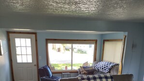 Ein Wohnzimmer mit grauem Sofa, einem Holztisch und einem blauen Sessel. Ein großes Fenster bietet Blick auf einen Garten und eine Tür mit Fußmatte.