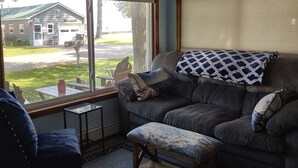 Ein Wohnzimmer mit einem dunkelgrauen Sofa, einem passenden Hocker und einem kleinen Tisch mit einer Vase. Durch ein Fenster ist ein Haus und ein Rasen zu sehen.