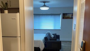 Un pasillo estrecho que conduce a un dormitorio con cama, ventilador de techo y ventana con persianas.