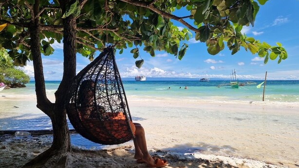 A beach scene with a hammock hanging from a tree, a clear blue sky, and boats in the water.
