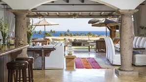A beachside lounge with a view of the ocean, featuring a bar, seating area, and a stone column structure.