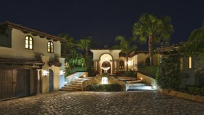 A courtyard with a stone pathway leading to a central fountain, surrounded by palm trees and a building with arched windows.