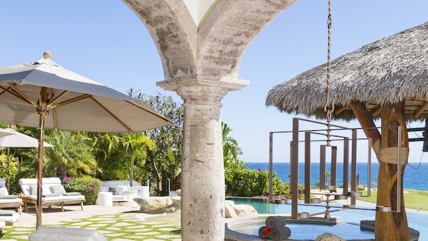 A poolside area with lounge chairs, a wooden table, and a thatched structure.