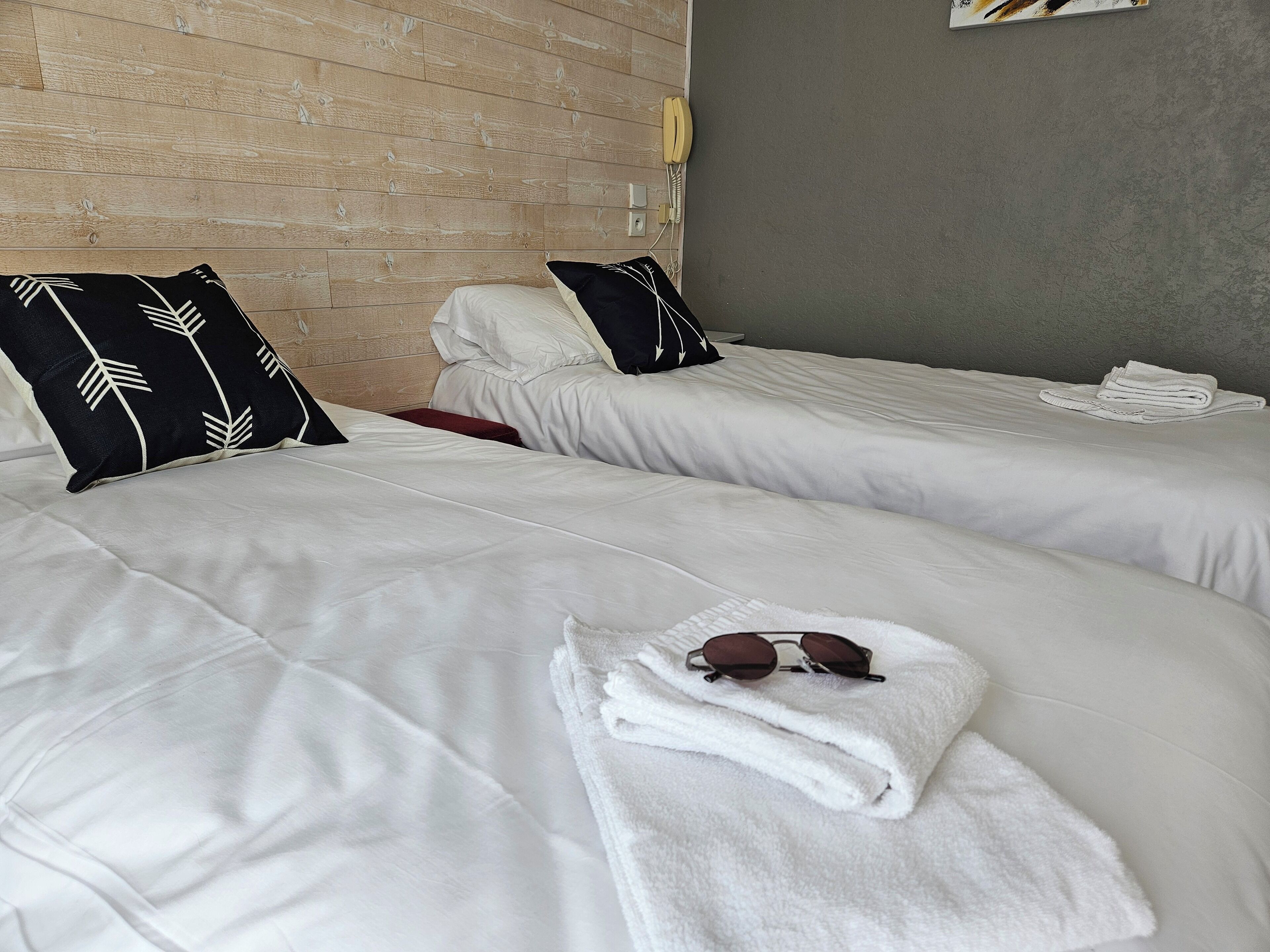 Two single beds with white linens, black pillows with white leaf patterns, a red bench, a white towel with sunglasses, and a telephone on the wall.