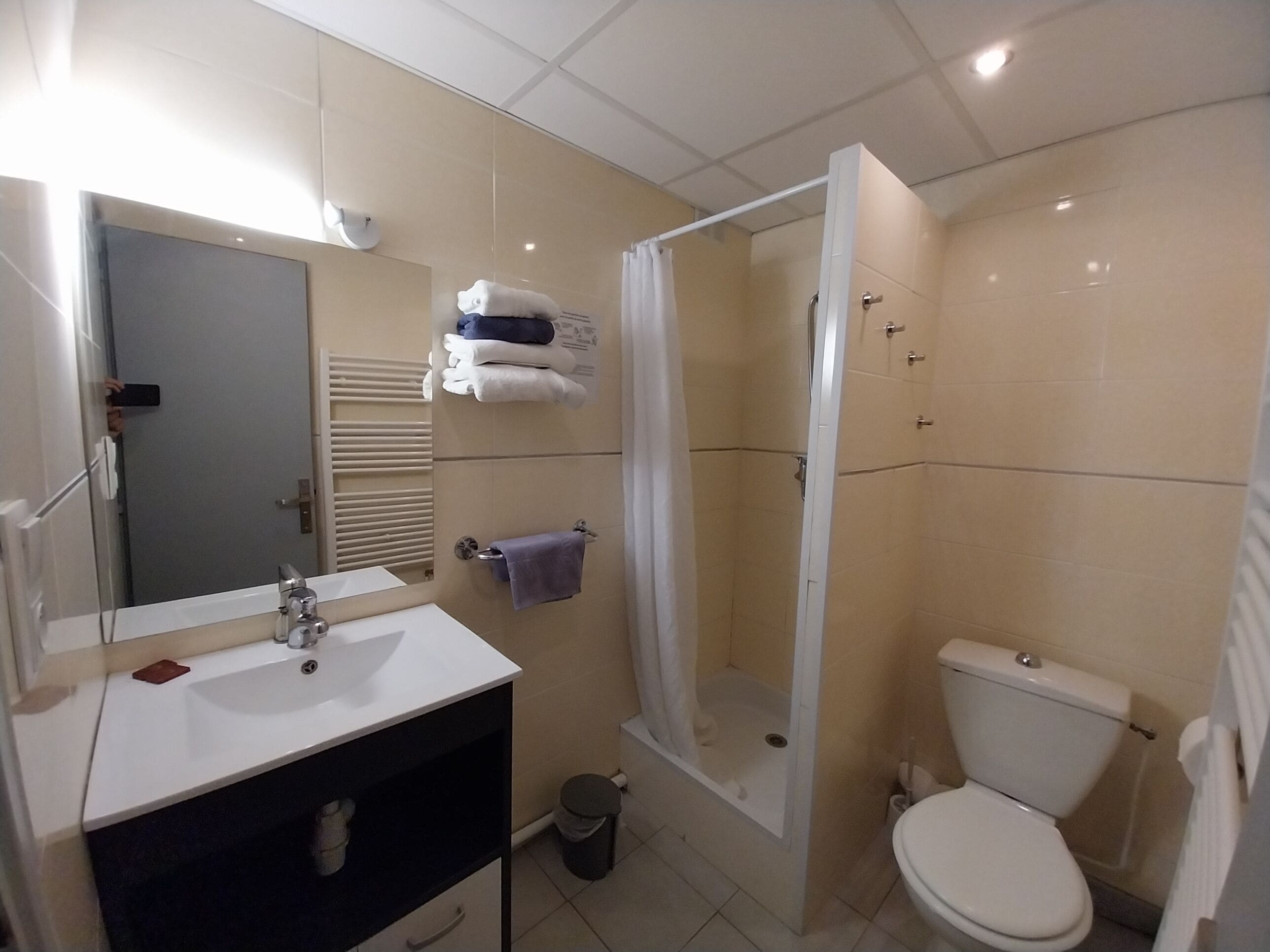 Un bany amb un lavabo blanc, un mirall i un vàter. Hi ha una zona de dutxa amb porta de vidre. Toletes penjades en un esparavell.