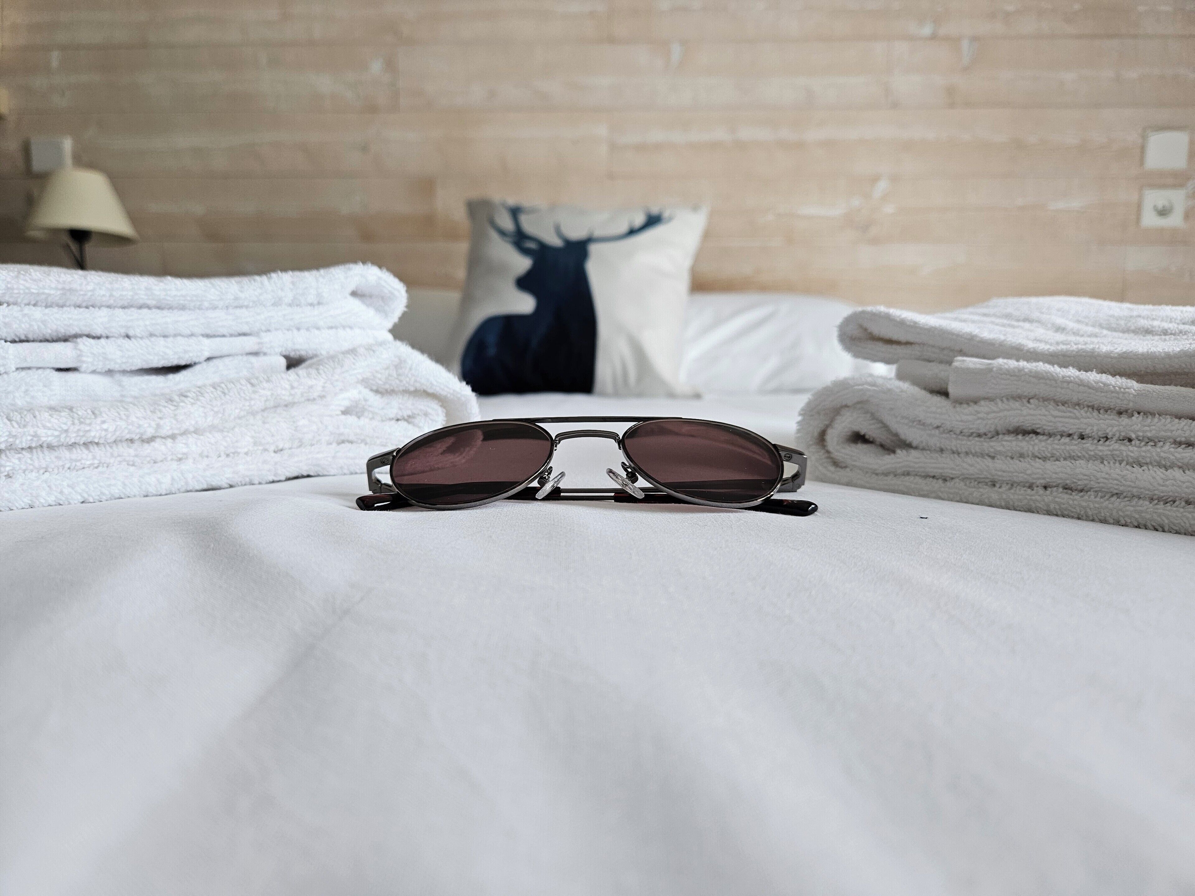 Una cama con sábanas blancas, un almohadón con un diseño de ciervo y un par de gafas de sol sobre la cama.