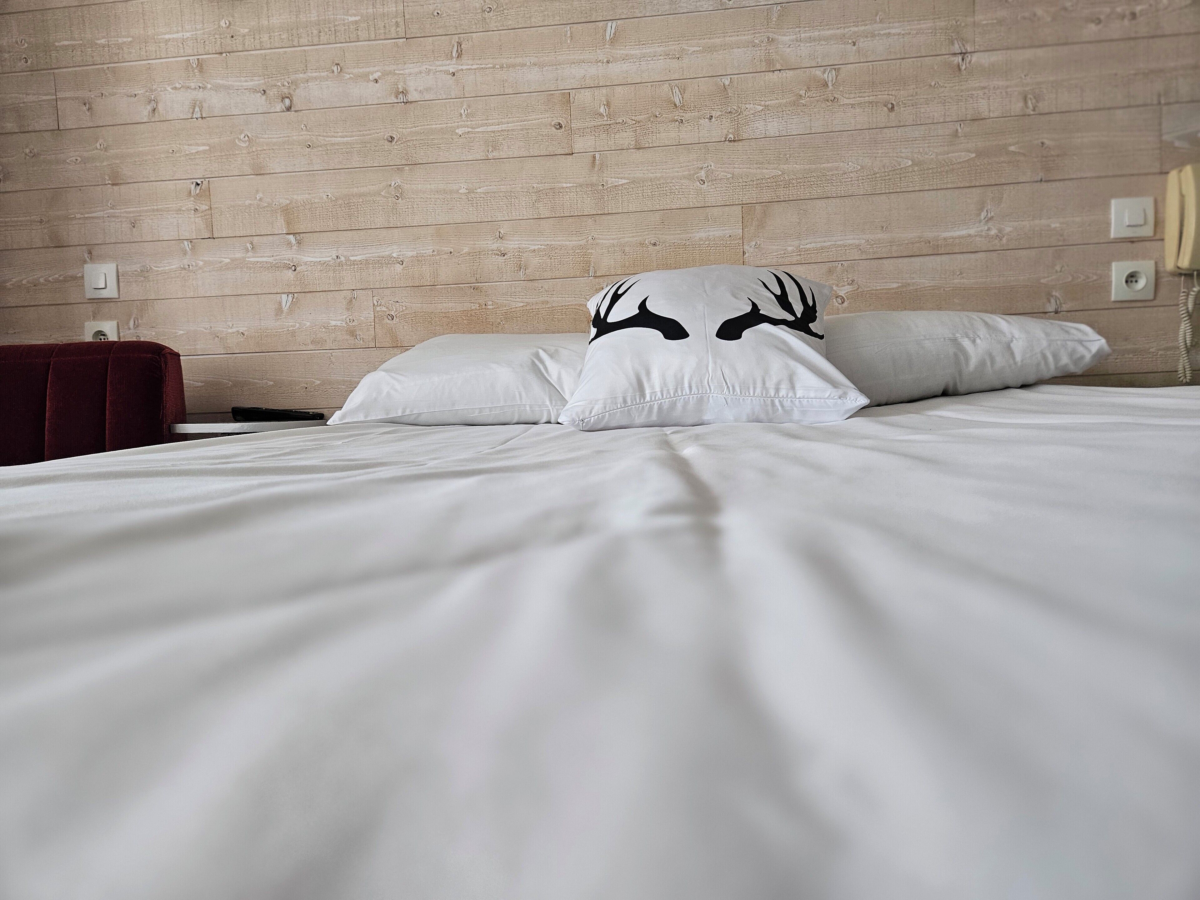 Una cama con ropa de cama blanca y dos almohadas, una con un diseño de ciervo, en una habitación con paneles de madera y una silla roja.