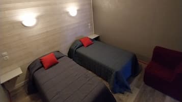 A room with two beds, each with a red pillow, a small table, and a red chair. There are wall-mounted lights and a vent on the wall.