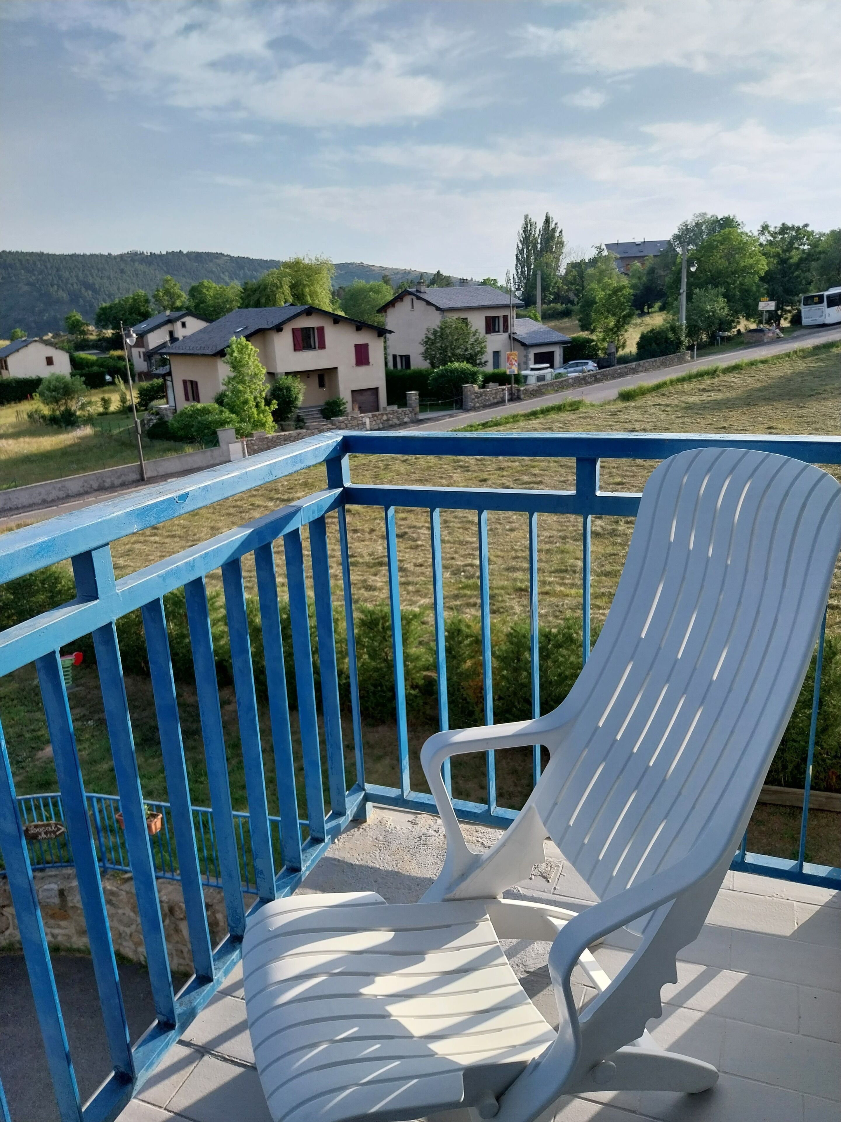 Een witte plastic stoel op een balkon met een blauwe reling, met uitzicht op een woongebied met huizen en groen.