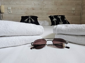 A neatly folded white towel, a pair of sunglasses, and two pillows with a feather pattern on a bed in a room with wooden walls.