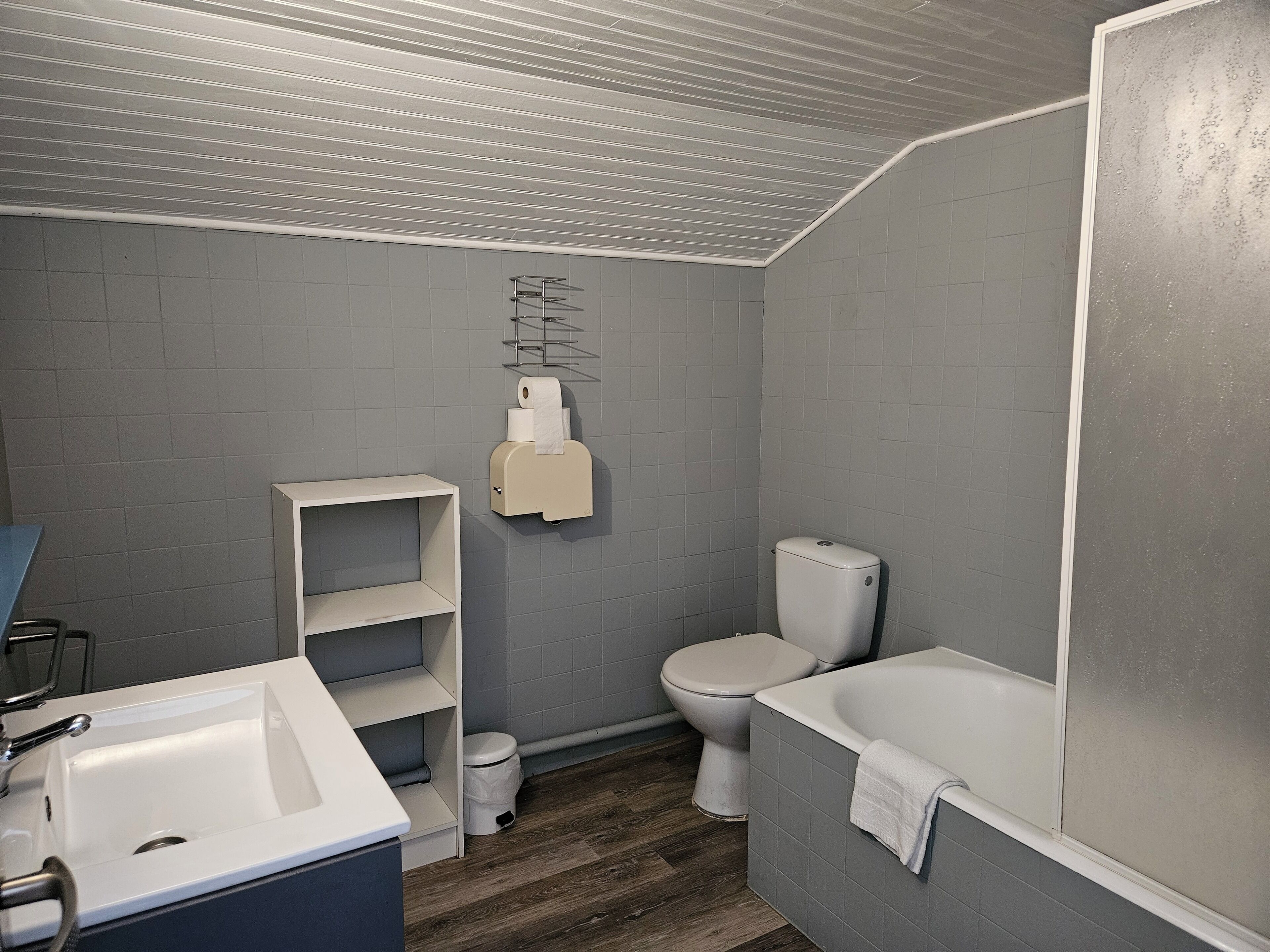 Un baño con lavamanos, inodoro y bañera blancos. Hay un mueble estante y un dispensador de toallas de papel en la pared.