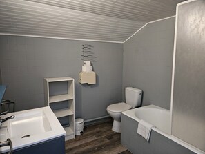 A bathroom with a white sink, toilet, and bathtub. There is a shelf unit and a paper towel dispenser on the wall.