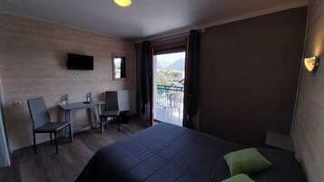 A hotel room with a bed, a desk, a chair, a TV, and a balcony with a view of the mountains.