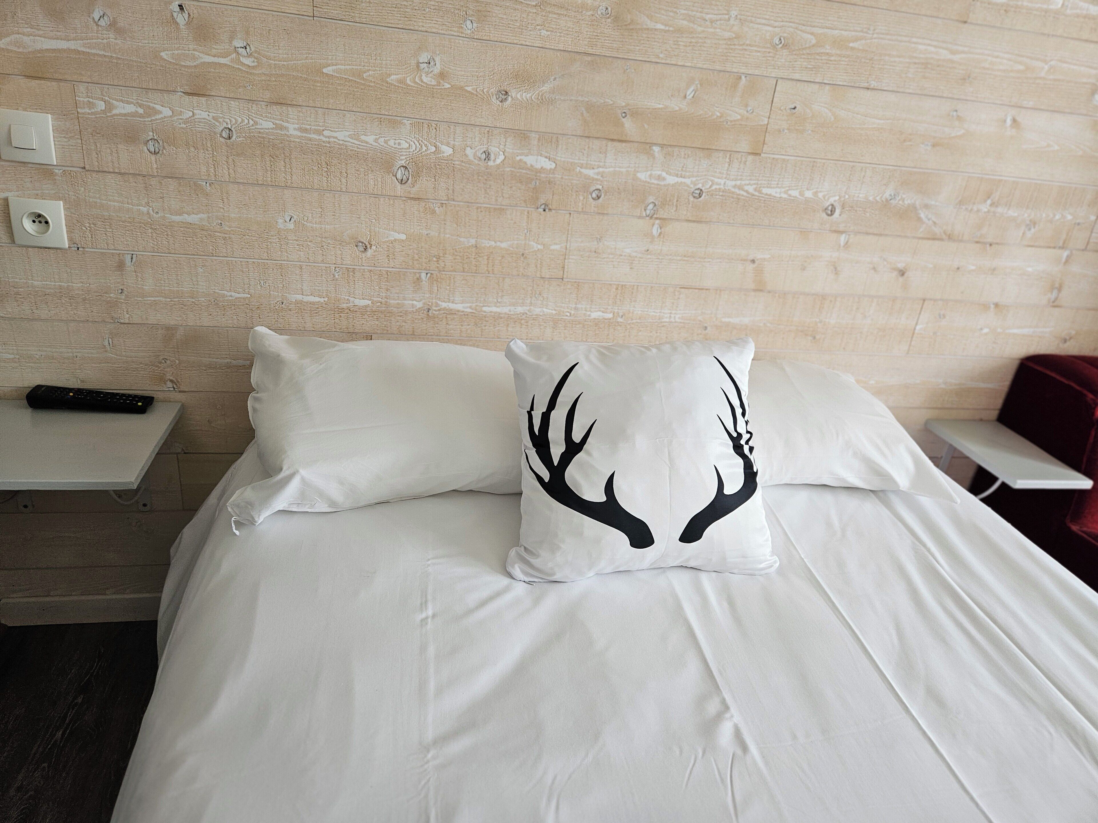A bed with white bedding and a deer antler pillow, a remote control on a bedside table, and a wooden headboard.