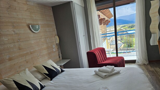 Habitación de hotel con una cama grande, una silla roja, una mesita pequeña y vistas a las montañas a través de la ventana.