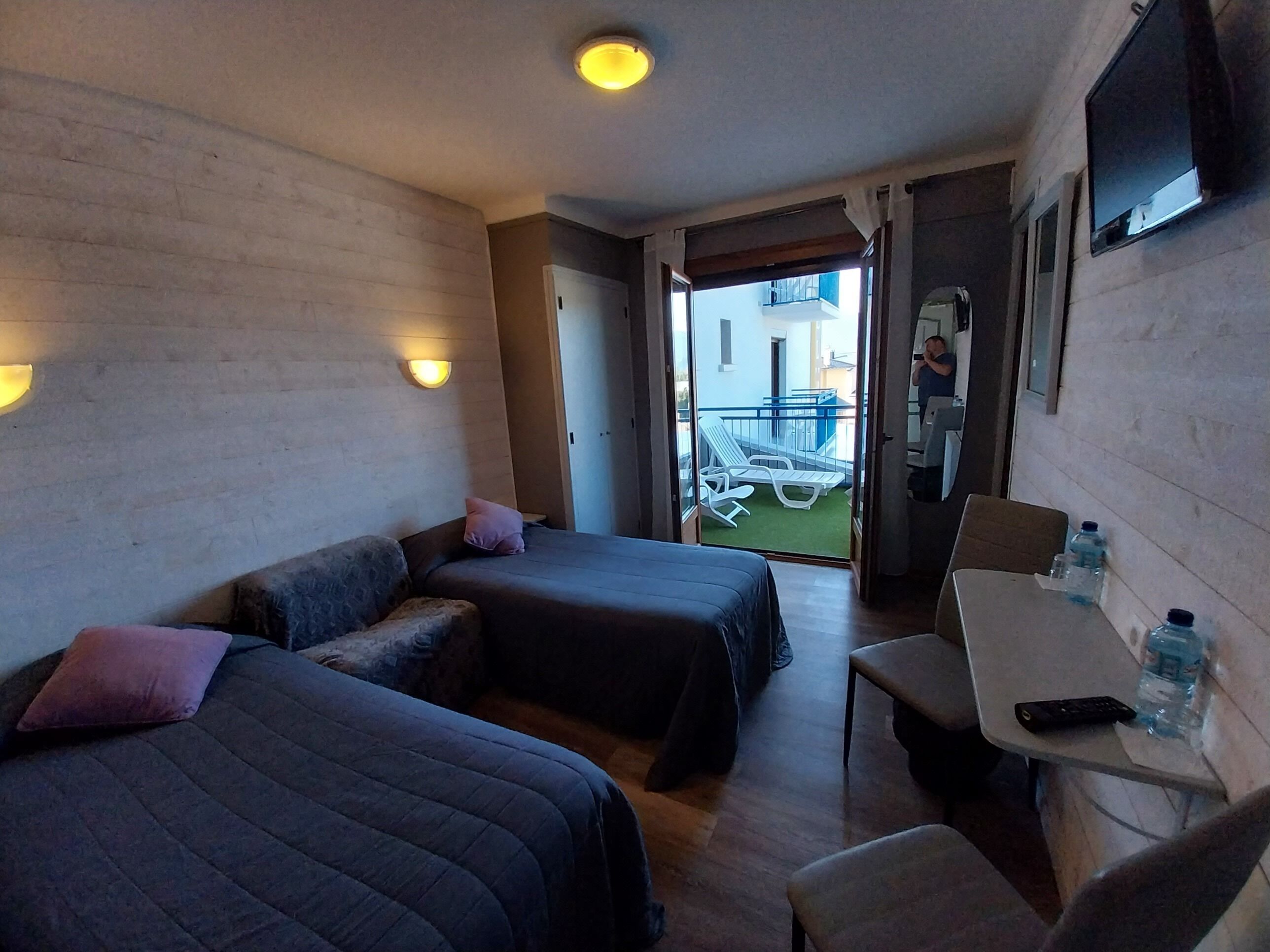 Une chambre d’hôtel compacte, équipée de deux lits, d’une petite table et d’une chaise. Elle dispose également d’un balcon avec vue sur l’extérieur.