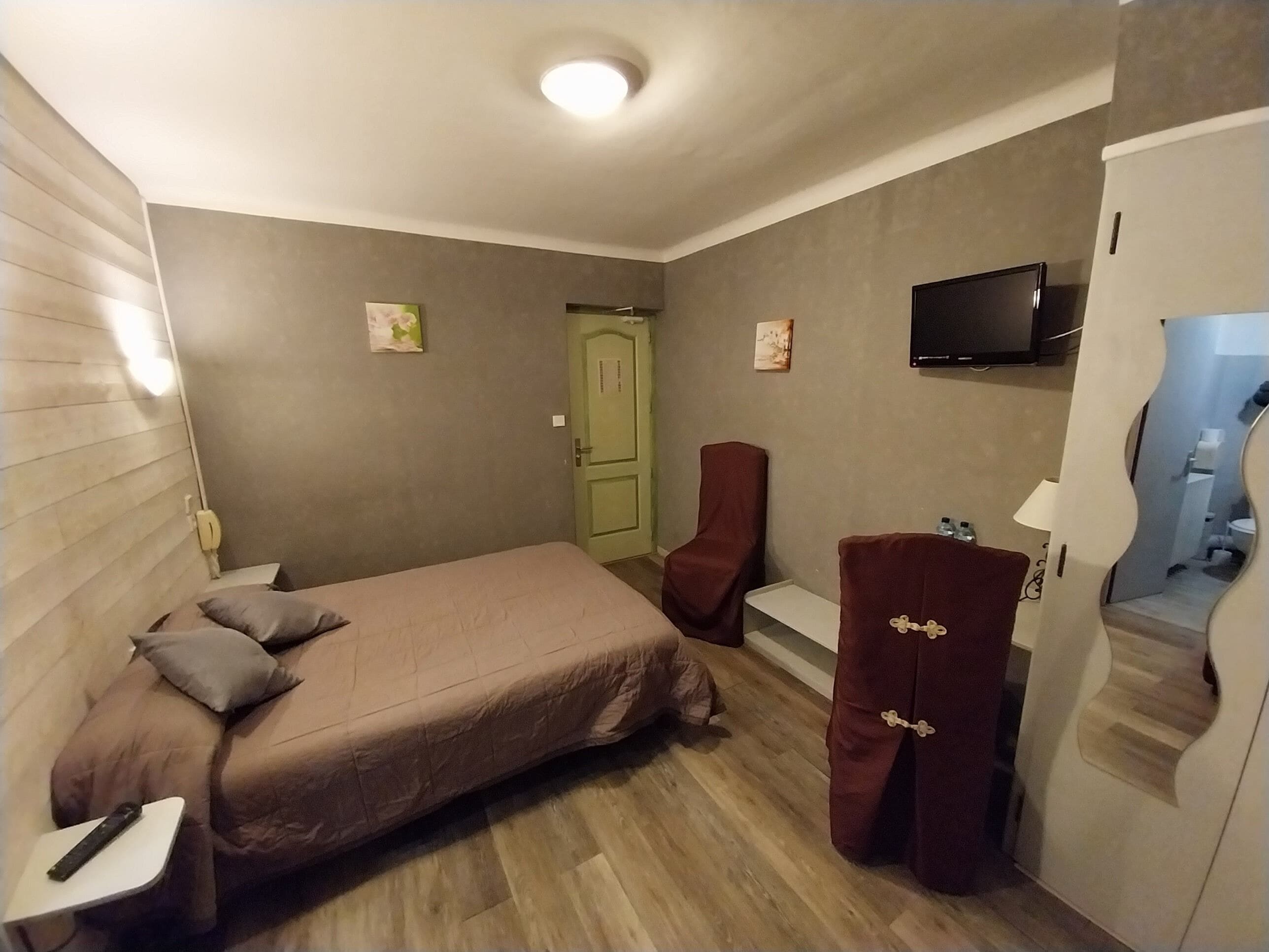 Une chambre d’hôtel de taille réduite, comprenant un lit simple, un petit bureau, une chaise, un téléviseur et une salle de bain visible à travers un miroir.