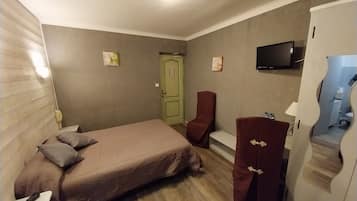 A compact hotel room with a single bed, a small desk, a chair, a TV, and a bathroom visible through a mirror.