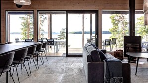 A modern interior with a large sliding glass door leading to a lakeside view, a seating area with a sofa and chairs, and a bar area with a grill. - Saunamäki Resort - Seafront Luxury (Salo)
