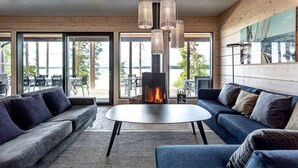 A modern living room with a fireplace, wooden walls, and large windows overlooking a deck. - Saunamäki Resort - Seafront Luxury (Salo)