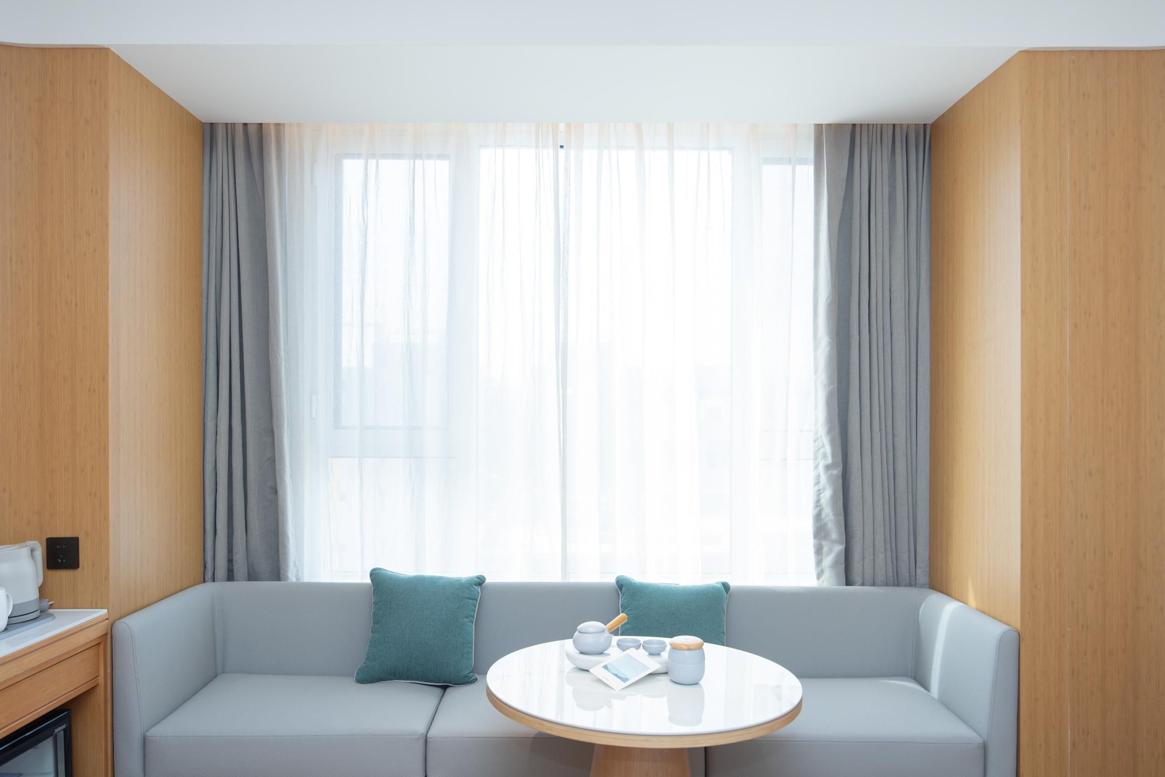 A modern hotel room with a grey sofa, a round table with a teapot and cups, and sheer curtains.