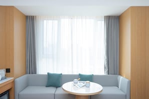 Een moderne hotelkamer met een grijze bank, een ronde tafel met theepot en kopjes, en dunne gordijnen.