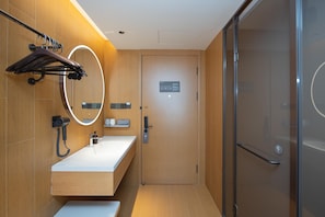 Een moderne badkamer met een houten wastafelmeubel, een spiegel met verlichting, een handdoekrek, een douchegedeelte en een deur met een bordje.