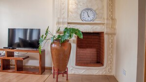 Ein Wohnzimmer mit Fernseher, einem großen Terrakottatopf mit Pflanze und einem Kamin aus Ziegelsteinen, über dem sich eine Uhr befindet.