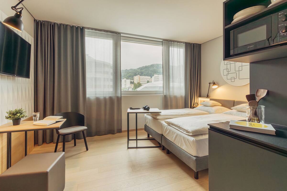 A hotel room with a bed, a desk with a chair, a TV, and a window with curtains.
