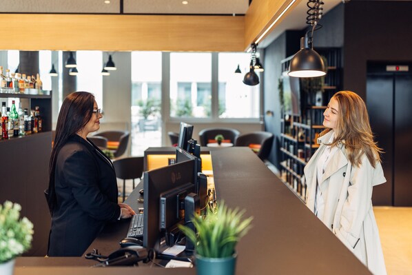 A reception desk with a computer and a telephone, a potted plant, and a bar area with shelves of bottles in the background. - Harry's Home Zürich Limmattal (Spreitenbach)