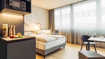 Ein modernes Hotelzimmer mit Bett, Schreibtisch mit Mikrowelle, Laptop, Esstisch und Blick auf die Stadt durch große Fenster.