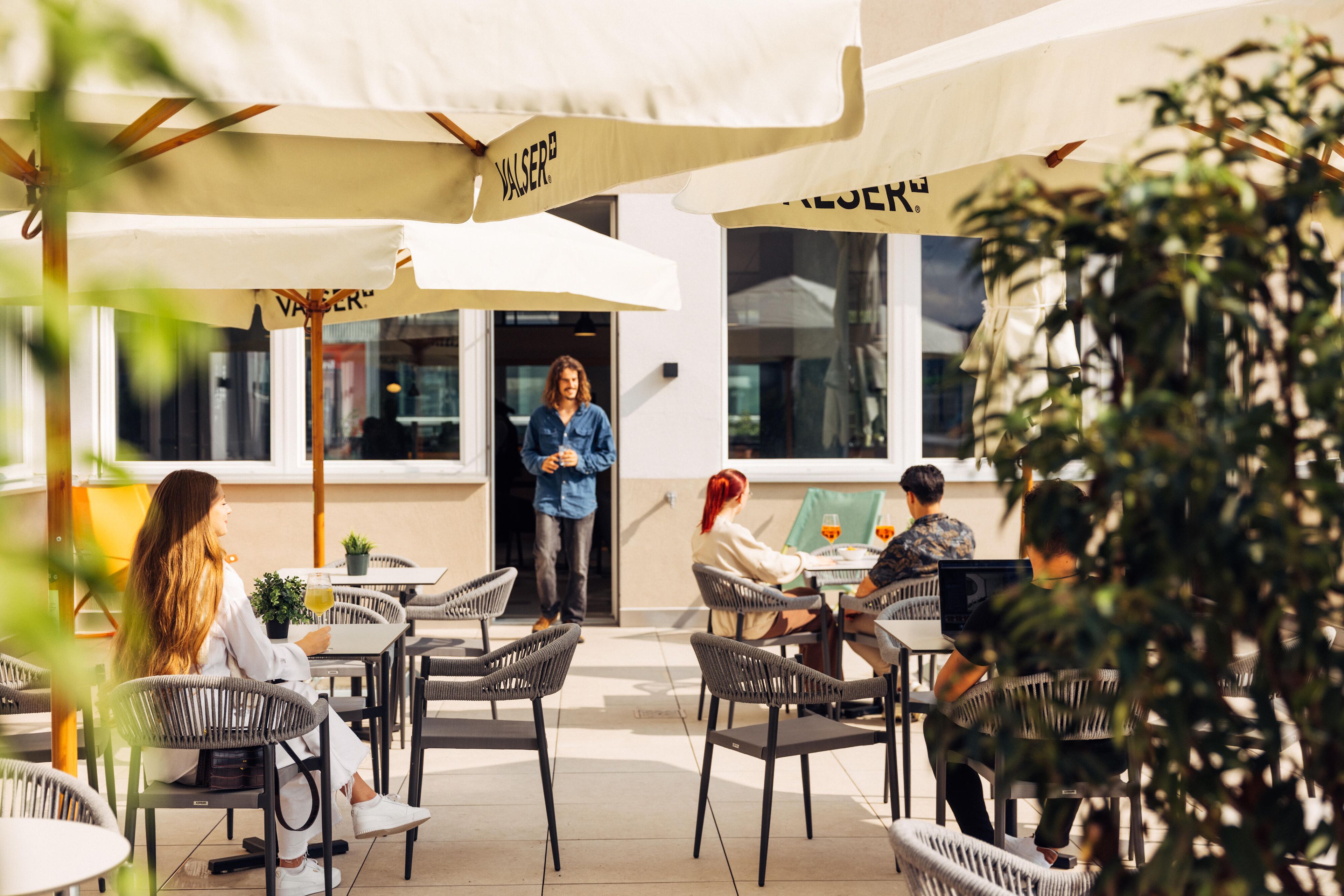 An outdoor café with wicker chairs and tables, umbrellas, and a person standing by the entrance.