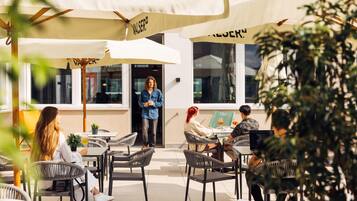An outdoor café with wicker chairs and tables, umbrellas, and a person standing by the entrance.