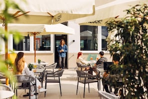 An outdoor café with wicker chairs and tables, umbrellas, and a person standing by the entrance. - Harry's Home Zürich Limmattal (Spreitenbach)