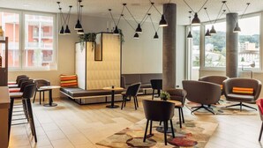 A modern lounge area with a sofa, chairs, and tables. There are large windows providing a view of the outside. - Harry's Home Zürich Limmattal (Spreitenbach)