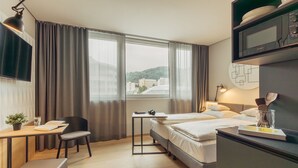 A hotel room with a bed, a desk with a chair, a TV, and a window with curtains. - Harry's Home Zürich Limmattal (Spreitenbach)