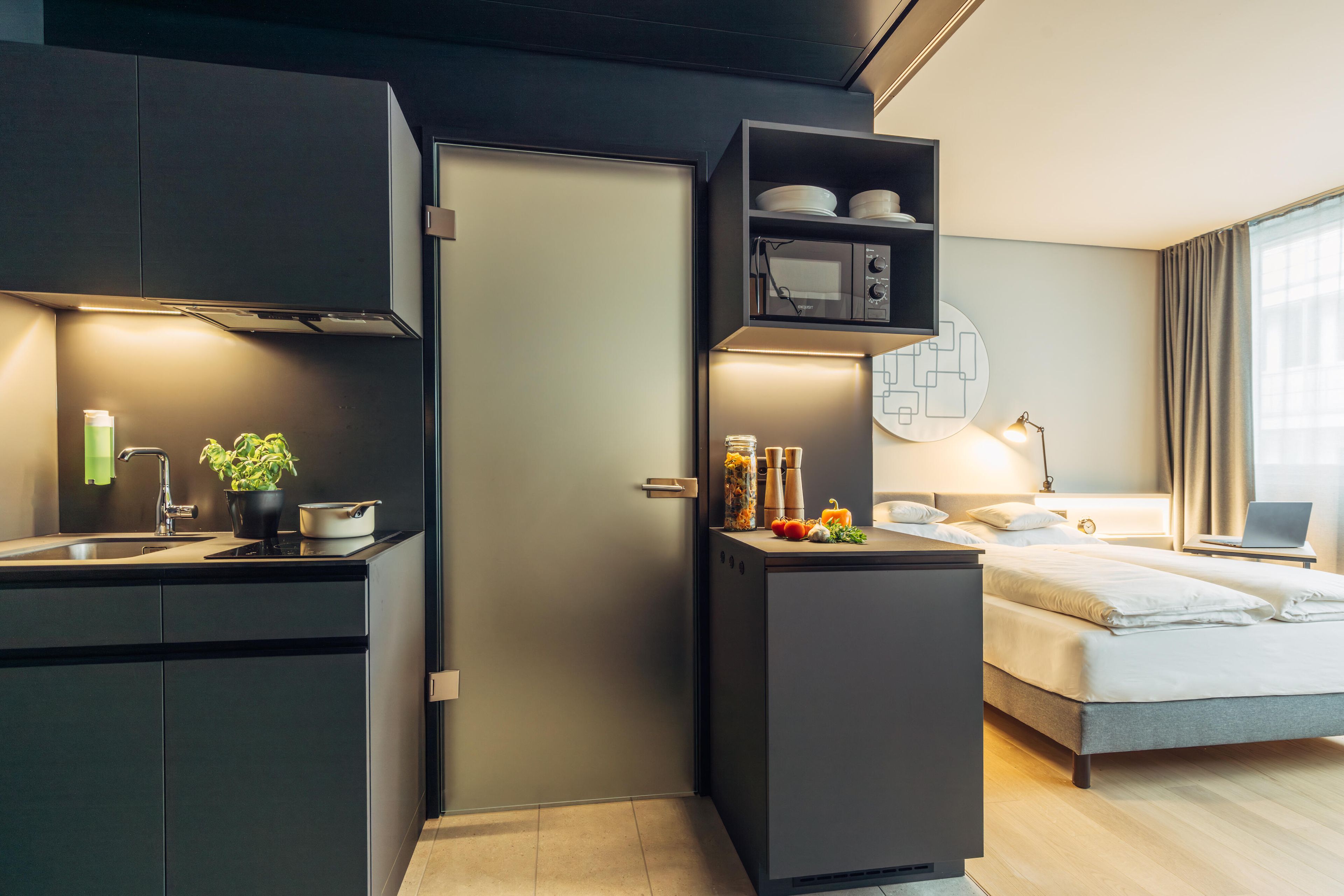 Una habitación de hotel compacta con kitchenette, una cama, un escritorio con una computadora portátil y una ventana con cortinas.