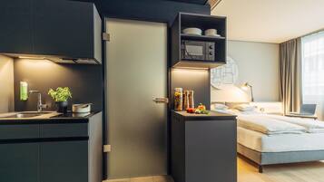 Una habitación de hotel compacta con kitchenette, una cama, un escritorio con una computadora portátil y una ventana con cortinas.