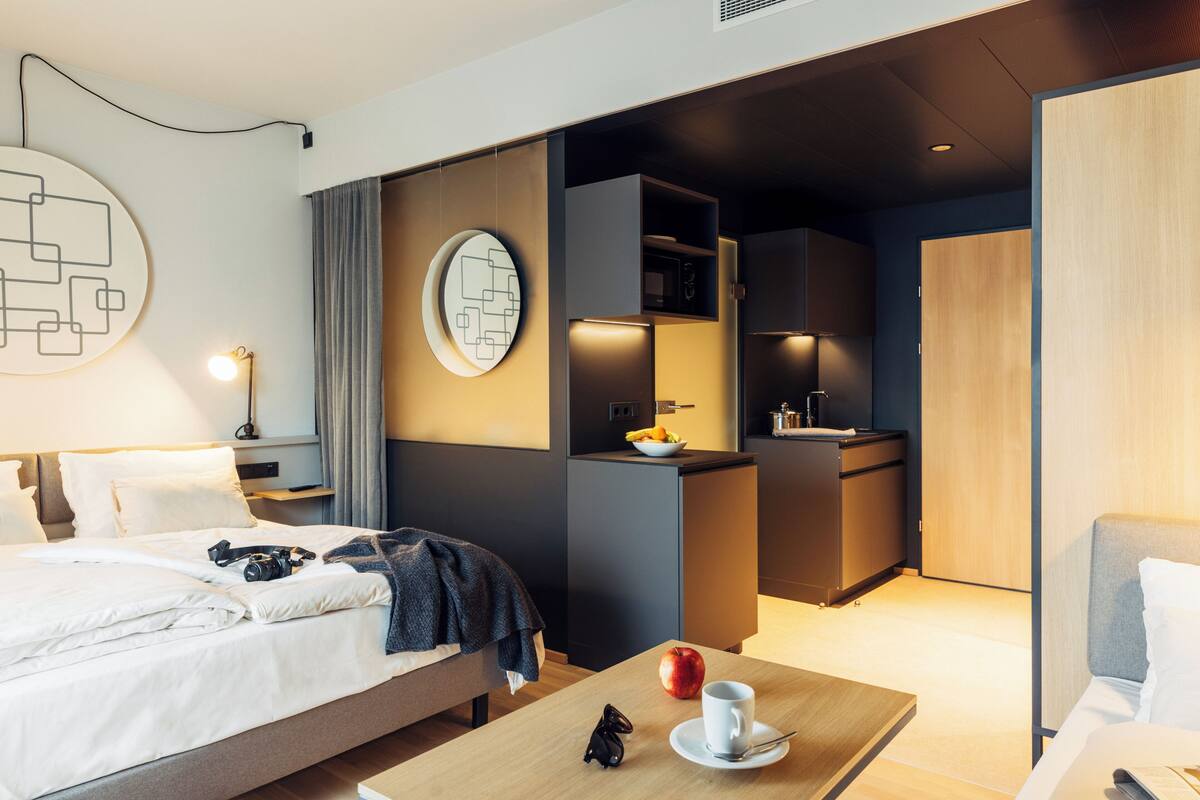 A modern hotel room with a bed, a desk, a small kitchenette, and a bathroom.