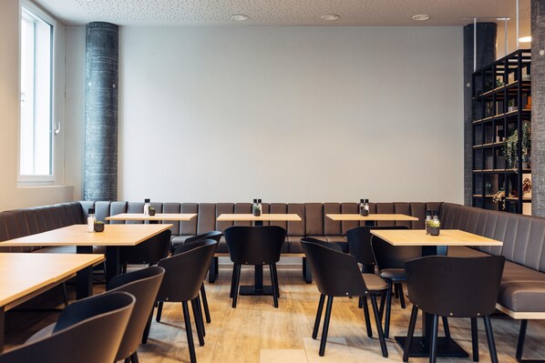 A modern restaurant with booths and tables, wooden flooring, and a bookshelf with plants. - Harry's Home Zürich Limmattal (Spreitenbach)
