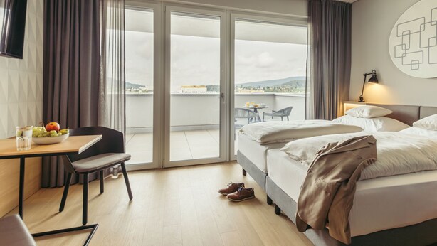 A hotel room with a bed, a desk with a chair, a dining table with chairs, and a view of the city through the window. - Harry's Home Zürich Limmattal (Spreitenbach)