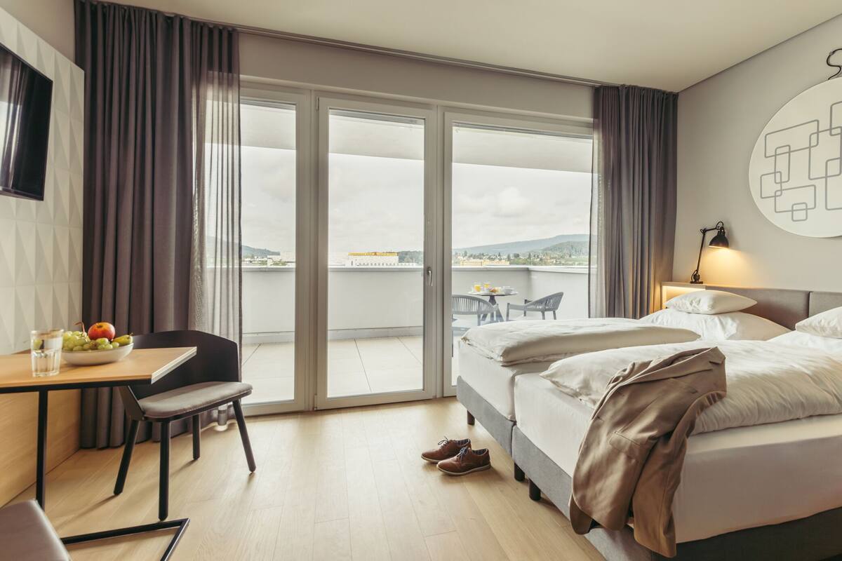 A hotel room with a bed, a desk with a chair, a dining table with chairs, and a view of the city through the window.