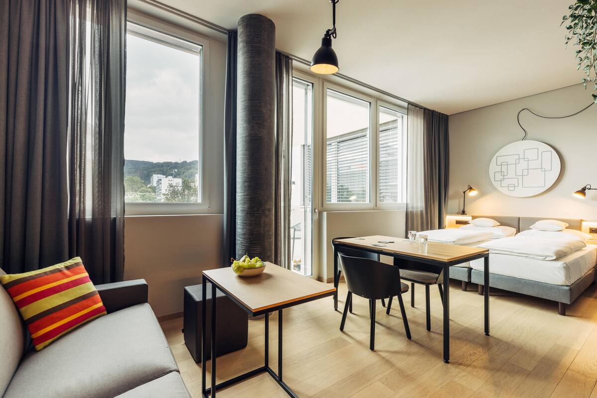 A modern hotel room with a sofa, a dining table with chairs, a bed, and a large window with curtains.