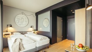A modern hotel room with a double bed, bedside tables with lamps, a fruit bowl on a side table, and a mirror on the wall. - Harry's Home Zürich Limmattal (Spreitenbach)