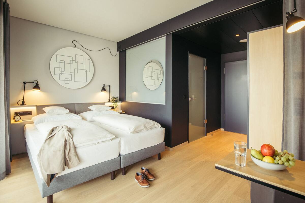 A modern hotel room with a double bed, bedside tables with lamps, a fruit bowl on a side table, and a mirror on the wall.