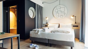 A modern hotel room with a bed, bedside tables, a desk, and a shower area. - Harry's Home Zürich Limmattal (Spreitenbach)
