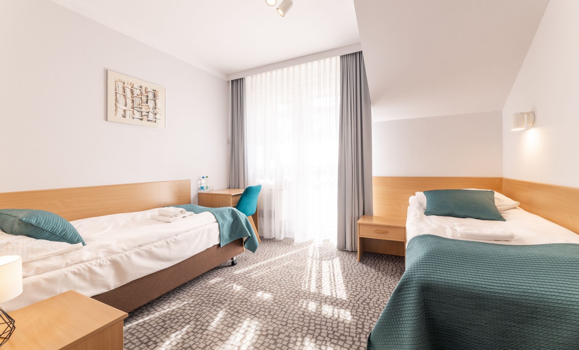 A hotel room with two beds, a desk, a chair, a window with curtains, and a wall-mounted artwork.